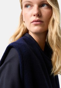 Námořnický fleecový plášť s volným designem, který má měkkou texturu a detailní záběr na rameno ženy a její vlasy na neutrálním pozadí.