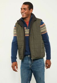 Gefütterte olivgrüne Weste mit einem Lammfellkragen, getragen über einem dunkelblauen Pullover mit mehrfarbigen Streifen und blauen Jeans.