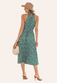 Vestito verde con scollo a halter, con motivo a macchie bianche, di lunghezza media, in tessuto leggero, indossato con un cappello di paglia e una borsa intrecciata.