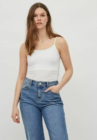 Top de tirantes finos ajustado blanco, combinado con unos vaqueros de tiro alto azules que presentan un cierre de botones y bolsillos. Textura de tela suave.