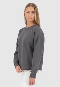 Felpa grigia con scollatura rotonda, maniche raglan e polsini a costine. Tessuto morbido, vestibilità rilassata, abbinata a pantaloni chiari.