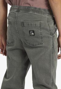 Los pantalones grises de cintura elástica cuentan con bolsillos traseros con un parche negro de logo. La tela tiene una textura suave con un ligero brillo.