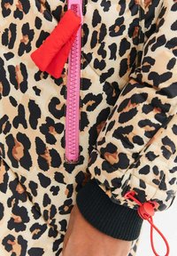 Pružná bunda v tygrovaném vzoru. Obsahuje růžový zip, červenou poutko a černé žebrované manžety s nastavovací šňůrkou.