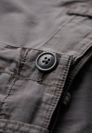 Grijs stof met een soepele textuur, voorzien van een zwarte knop met vijf gaten en het geperste 'Superdry' logo. Stiksel zichtbaar langs de naden.