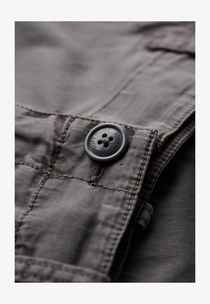 Grå stof med en glat tekstur, prydet med en sort knap med fem huller og præget "Superdry" logo. Syninger synlige langs sømmene.