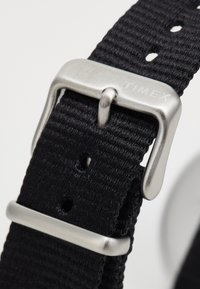 Cinturino da orologio in nylon intrecciato nero con fibbia in metallo argentato. Include fori regolabili per la dimensione e una incisione del marchio "TIMEX".