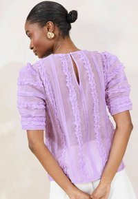 Blusa di chiffon lavanda con ruches verticali e dettaglio a chiave sul retro. Maniche corte a palloncino e una texture morbida.
