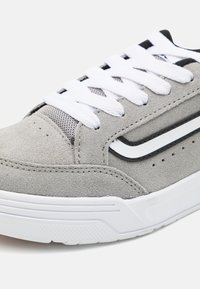 Grijze suède sneaker met witte veters, een mesh-tong en zwarte accenten. Beschikt over een gestructureerde zool en een klassiek ontwerp met stikseldetails.