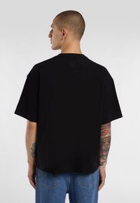 Black short-sleeve t-shirt with a relaxed fit, made of cotton, featuring a straight hem and minimalistic design. Visible tattoos on arm.
