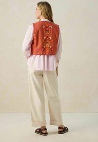 Un gilet brodé couleur rouille avec des motifs floraux, superposé sur un chemisier rose clair, associé à un pantalon large crème et des sandales marron.