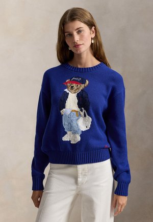 Maglione blu lavorato a maglia con grafica di un orso che indossa un cappello e una giacca rossi, con maglione bianco e jeans blu, polsini e orlo a coste.