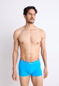 Blå bomulds boxershort med en hvid logo elastik, tætsiddende design og glat tekstur, vist på en model med en neutral baggrund.