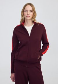 Femme portant une veste zippée bordeaux avec des rayures rouges sur les manches et un pantalon assorti, une chemise blanche en dessous, se tenant devant un fond uni.