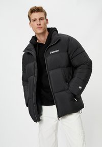 Zwarte pufferjack met ritssluiting aan de voorkant, uitgerust met een capuchon, doorgestikt textuur en logo op de borst. Draag het over een zwarte hoodie en witte broek.