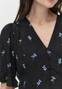 Schwarze Bluse mit blauem Blumenmuster, V-Ausschnitt, mit Stoff überzogene Knöpfe auf der Vorderseite und kurzen Puffärmeln mit Rüschensaum.