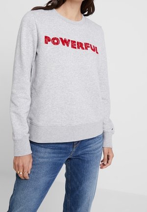 Vrouw draagt lichtgrijze trui met rode tekst "POWERFUL" over de borst en blauwe spijkerbroek, staande in een ontspannen houding.