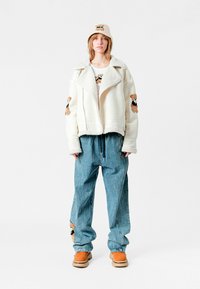 Chaqueta de borreguillo blanca con detalles bordados de oso, combinada con pantalones de mezclilla anchos de color azul claro, botas marrones y un gorro a juego con bordado de oso.