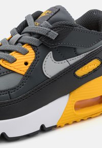Nike Air Max športni čevlji z temno sivo usnje, rumenimi in sivimi akcenti, teksturiranim jezikom ter vidno zračno blazino v belem podplatu.