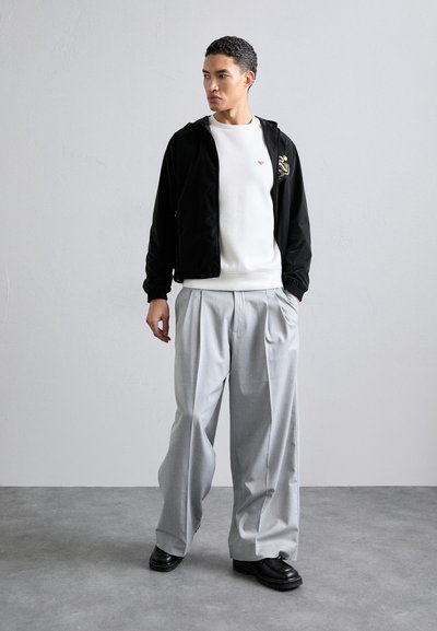 Casaco preto com fecho éclair e bordados em dourado, sweatshirts brancas e calças plissadas cinza claro. Sapatos pretos robustos completam o visual.