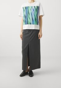 Camiseta blanca con un diseño abstracto colorido en verde y azul, combinada con una falda larga gris oscuro con una abertura frontal y zapatos negros.