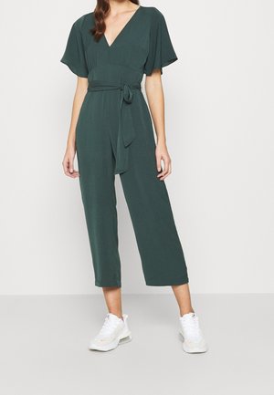 Femme portant une combinaison-pantalon vert foncé à jambes larges avec manches courtes et ceinture à nouer, associée à des baskets blanches, debout devant un fond uni.