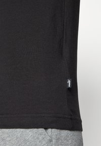 Melnā t-kreklā no viegla, gluda auduma ar noapaļotu apakšmalu un mazu logotipa birku sānos. Kombinēts ar gaiši pelēkiem sporta biksēm.