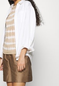 Bílá lehká košile s knoflíky přes béžový tie-dye top, doplněná hnědými šortkami. Má uvolněný střih a ohrnuté rukávy.