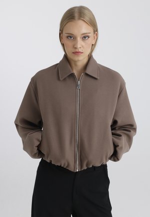 Jeune femme aux cheveux blonds et raides, portant une veste marron zippée et un pantalon noir, debout devant un fond clair uni.