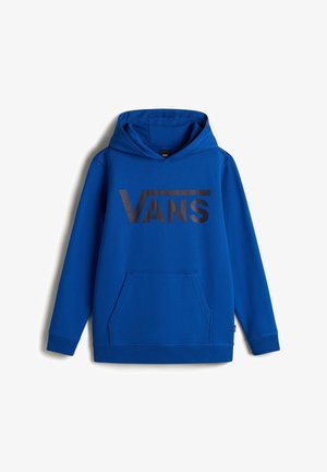 Personne portant un sweat à capuche bleu Vans avec les mains dans la poche kangourou, pantalon noir et baskets noires et blanches, debout sur un fond blanc.