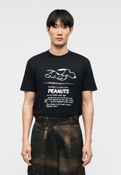 Νεαρός άνδρας που φοράει μαύρο T-shirt με λευκό γραφικό Snoopy και κείμενο που προωθεί τις κόμικς Peanuts, συνδυασμένο με σκούρο παντελόνι με μοτίβο.