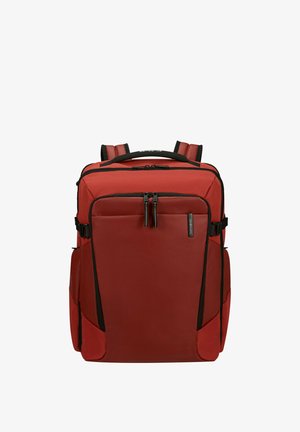 Zaino rettangolare rosso con spallacci imbottiti, tasca frontale con cerniera, tasche laterali e dettagli con cerniere nere, in piedi su sfondo bianco.