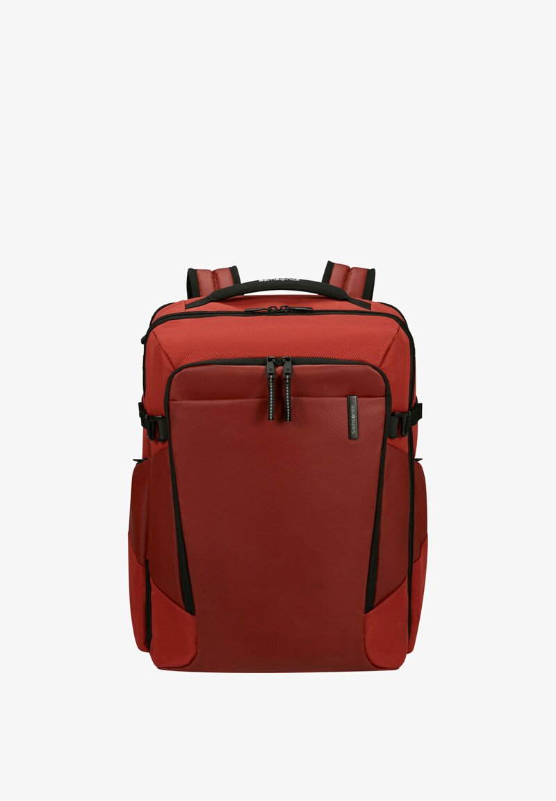 Zaino rettangolare rosso con spallacci imbottiti, tasca frontale con cerniera, tasche laterali e dettagli con cerniere nere, in piedi su sfondo bianco.