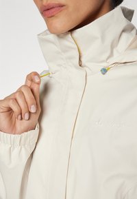 Beige vanntett jakke med høy krage, elastiske mansjetter og en gul pull-tab. Har en subtil logo og en glatt tekstur.