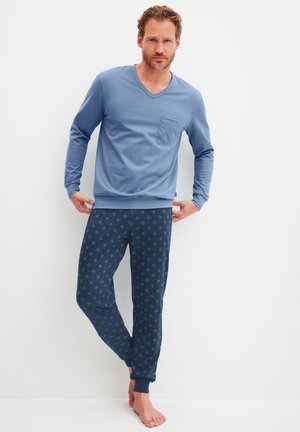 Man in een blauw, lang mouwig V-hals shirt en gemêleerde marineblauwe joggingbroek, blootsvoets staand tegen een witte muur.