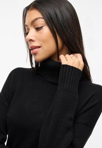 Svart polotröja i mjukt stickat material med ribbstickade muddar, med en figursydd silhuett och minimalistisk design.