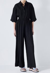 Combinaison noire avec col boutonné, manches courtes retroussées et taille élastique. Paire de sandales à motif visible aux pieds.