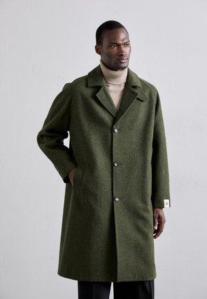 Zaļš vilnas mētelis ar izgriezumu, piecu pogu aizdari, sānu kabatām un birku kreisajā piedurknē. Modelis valkā beigspēļu turtleneck.