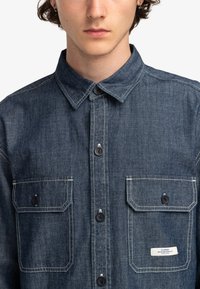 Denimskjorte i mørkeblå med knaplukninger, to brystlommer, kontrasterende hvid syning og et mærke på den venstre lomme.