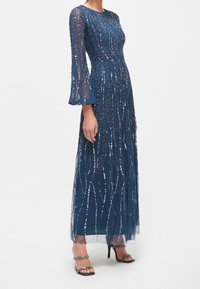 Lång marinblå klänning i genomskinligt tyg med intrikata pärlbroderier, med långa ärmar och en flödande kjol, kompletterad med klackar med remmar.