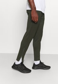 Olijfgroene taps toelopende sweatpants met ritsen bij de enkel, gecombineerd met zwarte sportschoenen en witte enkelsokken. Soepele stoftextuur.