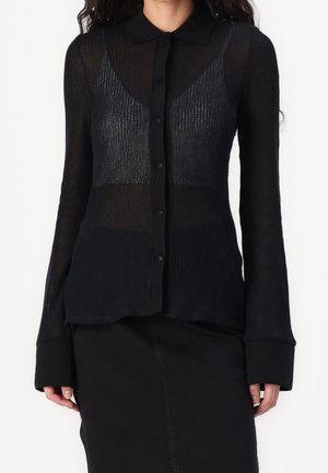 Kobieta ubrana w półprzezroczystą, czarną, zapinaną na guziki bluzkę z długim rękawem, na czarnym biustonoszu, zestawioną z czarną spódnicą na białym tle.