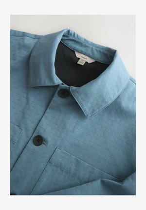 Blau strukturiertes Hemd mit einem Kragen, zwei schwarzen Knöpfen und einer Brusttasche. Das Etikett innen zeigt Größe M an.