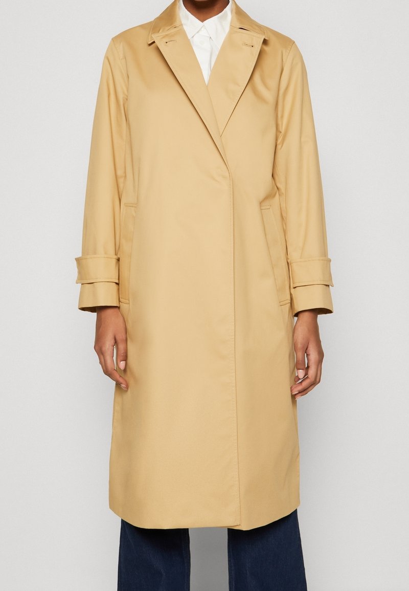 Beige trenchcoat med dobbeltspaltet design, lange ærmer og foldbare manchetter. Glat tekstur, mellemlang og med sidelommer.