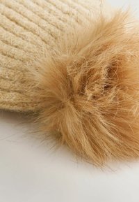 Caciula tricotată bej, cu un pom-pom mare din blană sintetică călduță pe vârf. Modelul texturat este proeminent și oferă un plus de adâncime designului.