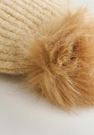 Chapeau en tricot beige avec un grand pompon en fausse fourrure tan duveteux sur le dessus. Le motif texturé est bien en évidence et apporte de la profondeur au design.