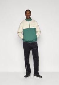 Πράσινο και μπεζ ζακέτα με φερμουάρ, μοτίβο με πλέγμα και λογότυπο, σε συνδυασμό με μαύρο παντελόνι κορντόι με λευκά γράμματα και κεντημένες λεπτομέρειες.