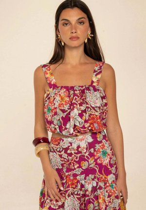 Mujer con un top morado sin mangas con flores y falda a juego, complementado con grandes pendientes dorados y dos pulseras gruesas.