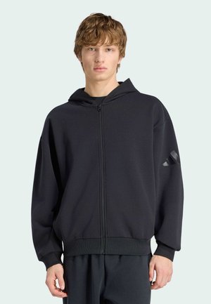 Mladý muž nosící černou mikinu na zip s jemným logem na levém rukávu, stojící před světlým jednobarevným pozadím.