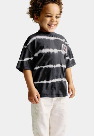 Maglietta nera tie-dye con strisce bianche, maniche corte e una piccola toppa grafica sul petto, abbinata a pantaloni di colore chiaro.