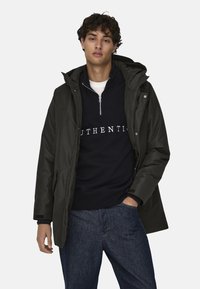 Sort parka med hætte, udstyret med lynlås foran og sidelommer, lagt over en mørk marineblå sweatshirt med hvid tekst "AUTHENTIC".